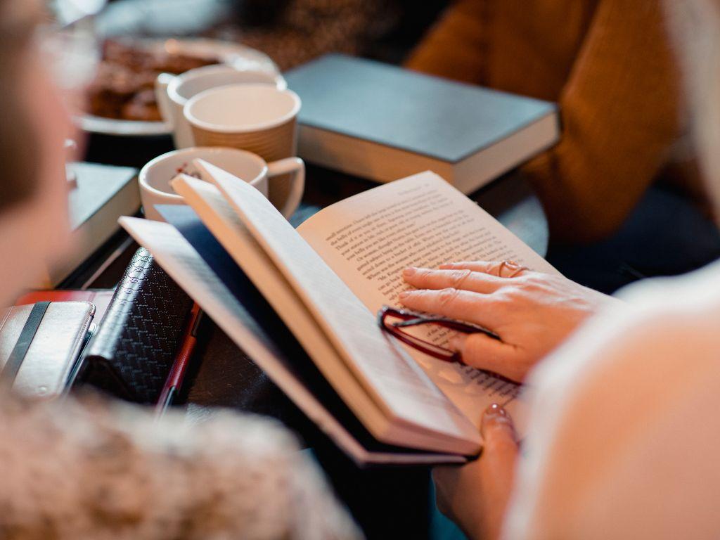 Imatge recurs on es veu una mà a sobre d'un llibre obert. De fons es veu una taula amb tasses de cafè